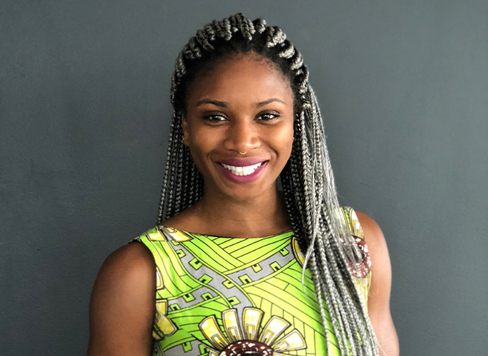 Bénédicte Bicaba, smiling to camera in a vivid green dress with floral pattern in front of a grey wall.