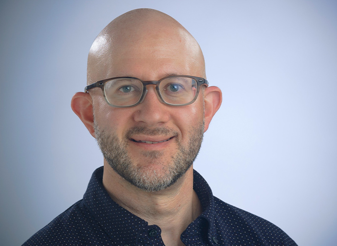Josh Belzman, wearing grey glasses, smiling in front of a grey background
