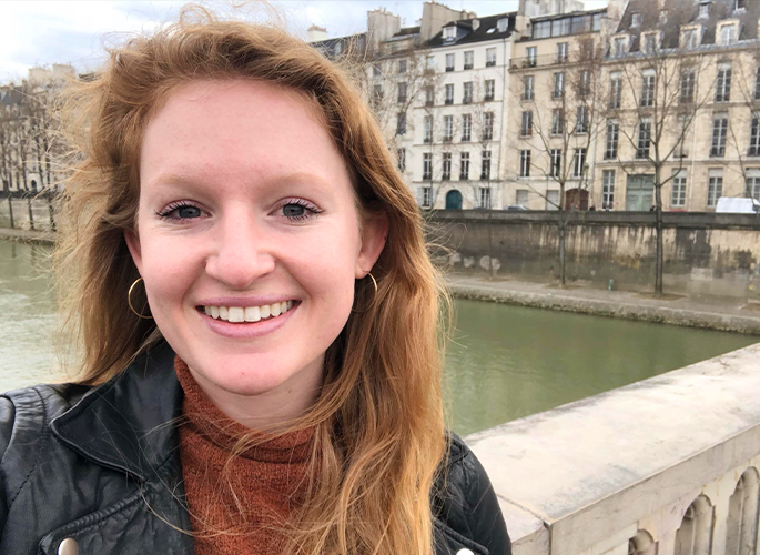 Megan Arnold in a dark jacket and loose hair standing on a bridge with a row of buildings in the background