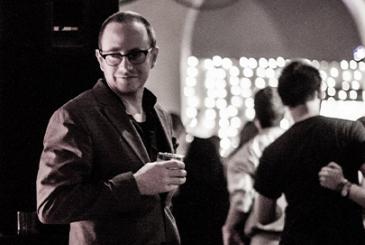 Michael Seguin standing next to a dance floor holding a tumbler. The photo is in black and white.