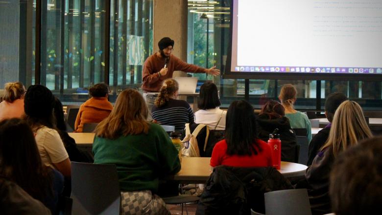 HUM 101 Student Harjot Singh at HUM 101 Final Presentations, Au '21 Freshman student Harjot Singh dynamically holds his left arm out towards a projector screen displaying his digital e-portfolio, watched by a group of his seated classmates.