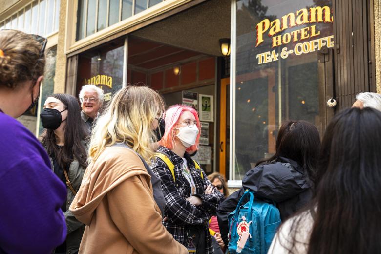 A mixed group of students, largely masked, stand talking in front of the Panama Hotel in Seattle' International District. A sign on the building window reads "Panama Hotel Tea & Coffee".