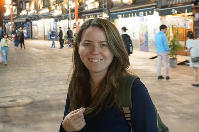 Maggie Le standing smiling in a shopping district street in the evening; people are milling around the background