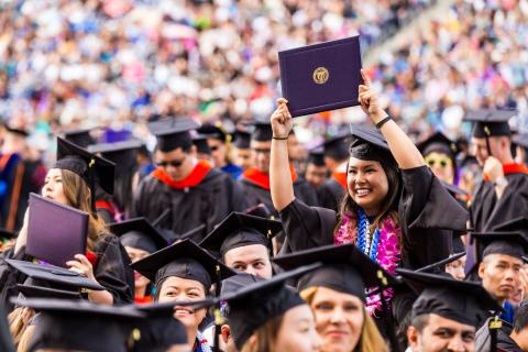 A large crowd of graduating students in caps and gowns, with one woman standing, smiling, and holding her diploma above her head