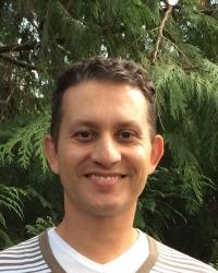 Eduardo Viana da Silva smiling to the camera in a striped shirt in front of a leafy green background