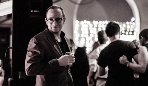 Michael Seguin standing next to a dance floor with dancers facing away from the camera. The photo is in black and white.