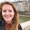 Megan Arnold in a dark jacket and loose hair standing on a bridge with a row of buildings in the background