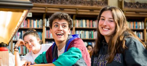 Three students smiling while seated at a shared library table. The lighting is warm and soft.