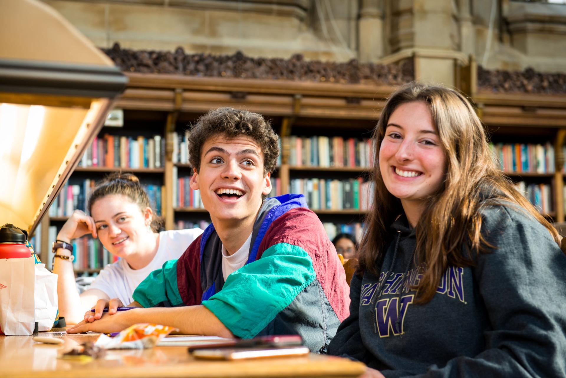 Three students smiling while seated at a shared library table. The lighting is warm and soft.